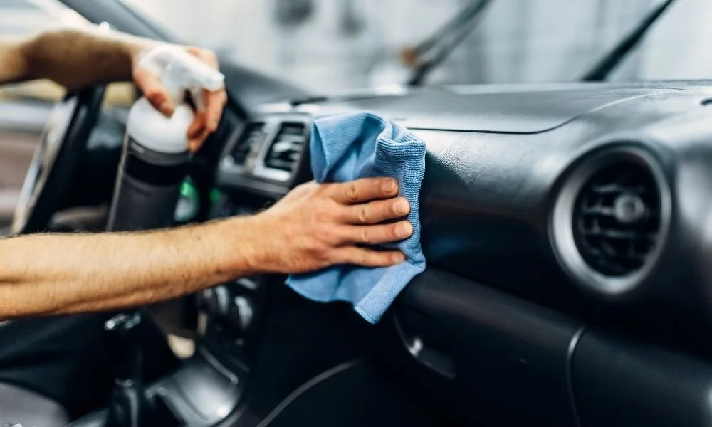 Person cleaning car dashboard with microfiber cloth and spray bottle, highlighting car detailing process and vehicle maintenance.