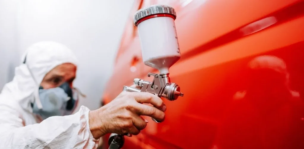 Person in protective gear applying ceramic coating to a red car using a spray gun, illustrating car detailing and maintenance services.