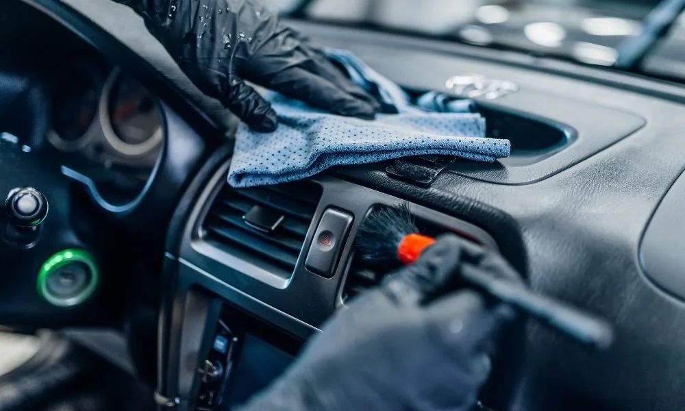 Hands wearing black gloves cleaning a car dashboard with a microfiber cloth and brush, highlighting car detailing preparation.