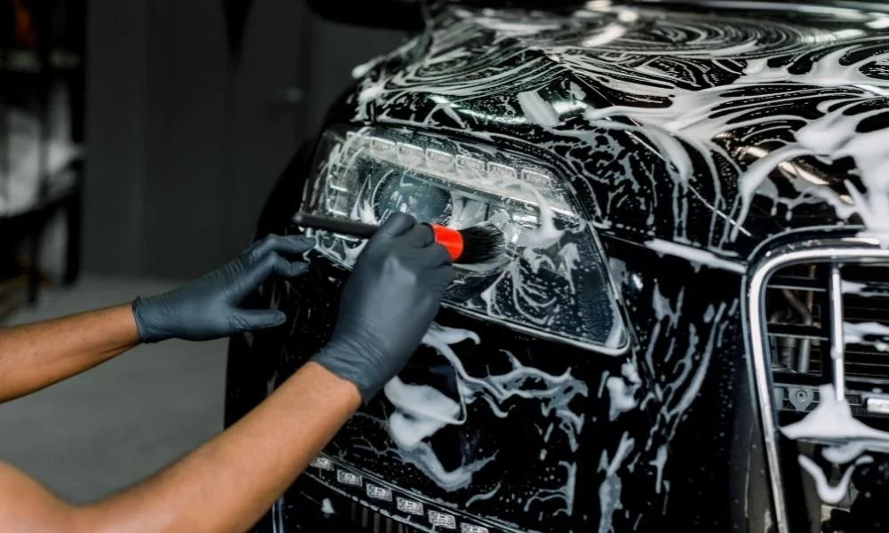 Person applying foam soap to the headlight of a black vehicle, emphasizing automotive detailing and preparation for ceramic coating.