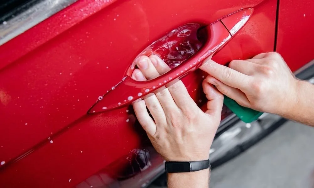 Hands applying automotive clear bra film to a red car door handle, demonstrating vehicle protection and detailing techniques.
