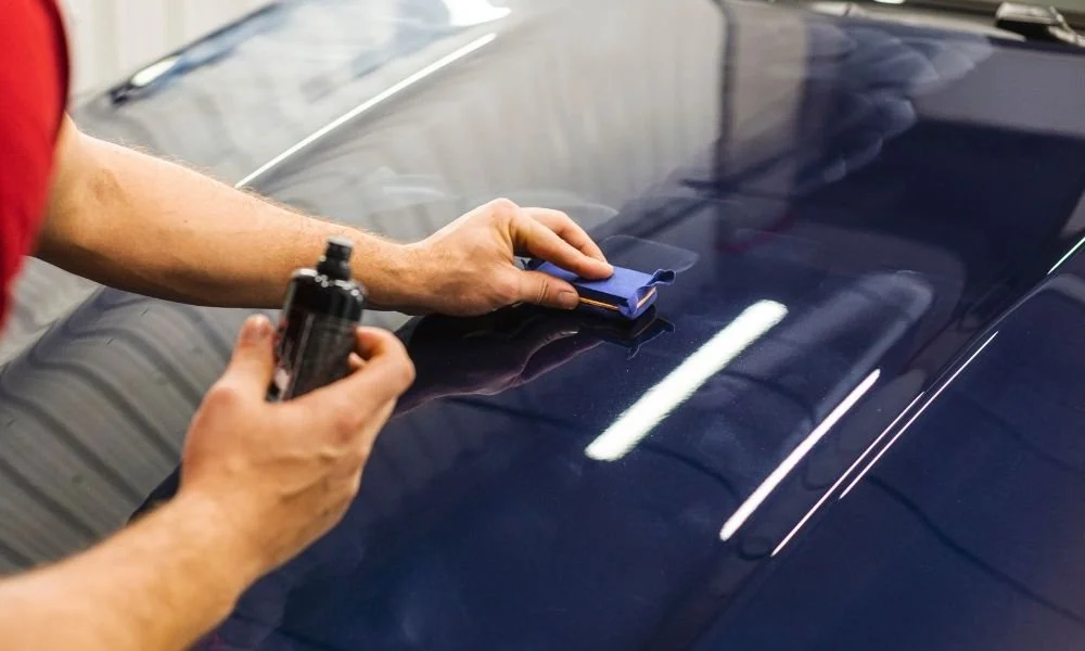 Person applying ceramic coating to a car's hood using a blue applicator pad, with a bottle of product in hand, demonstrating automotive detailing techniques.