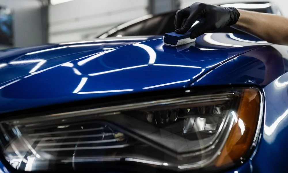 Person applying ceramic coating to a blue car's hood, showcasing vehicle detailing and paint protection techniques in a professional garage setting.