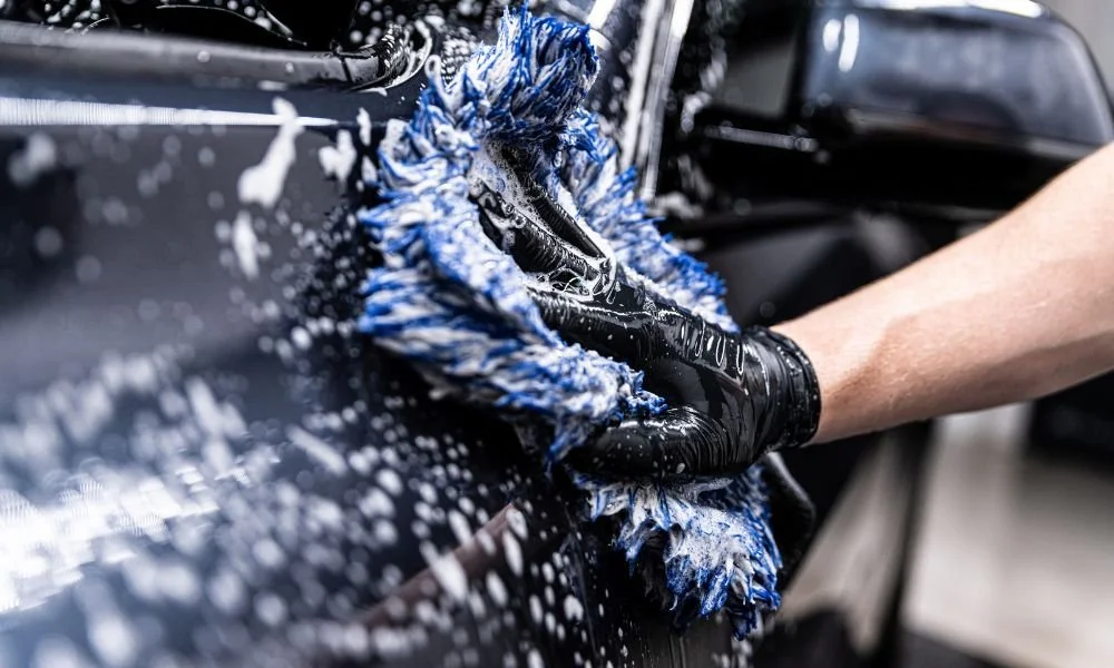 Person using a microfiber wash mitt to clean a car's surface with soapy water, emphasizing professional hand wash techniques for vehicle maintenance.