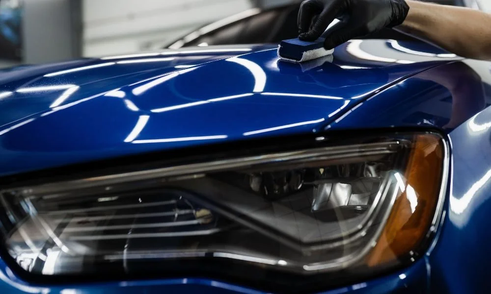Person applying car wax on a blue vehicle's hood, showcasing car detailing techniques to protect paint from environmental damage.