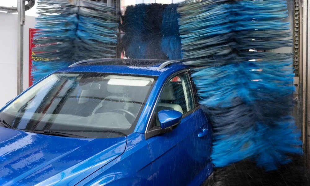 Blue car in automatic car wash with spinning brushes and water spray, illustrating common dangers of automatic car washes.