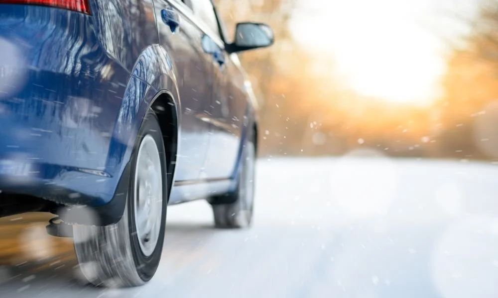 Blue car driving on snowy road with winter scenery, illustrating tips for protecting car exterior in winter weather.
