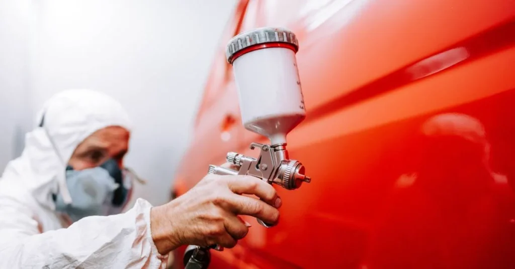 Person in protective gear using a spray gun to apply paint on a vehicle, emphasizing the importance of paint correction in car detailing.