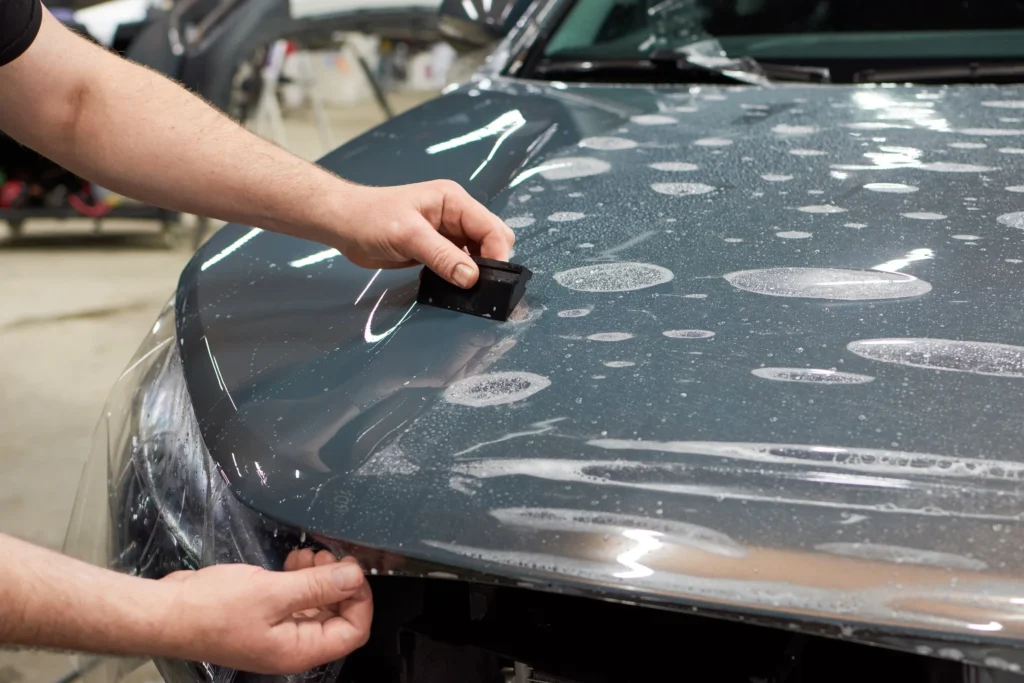 Person applying paint protection film (PPF) on a car hood, demonstrating vehicle protection against scratches and damage.