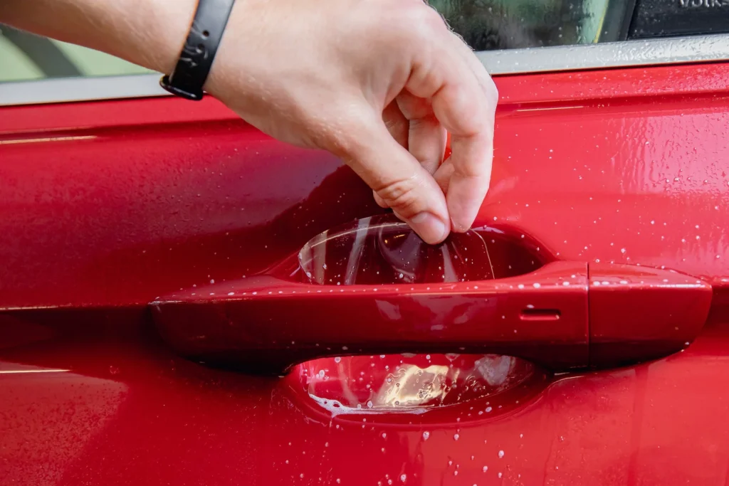 Hand applying paint protection film on a red car door handle, demonstrating car detailing technique for protecting vehicle surfaces.