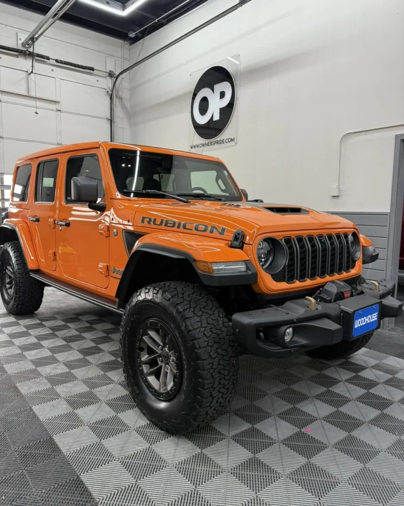 Orange Jeep Rubicon parked in an automotive detailing shop, showcasing paint protection film (PPF) installation, with Owner's Pride logo visible on the wall.