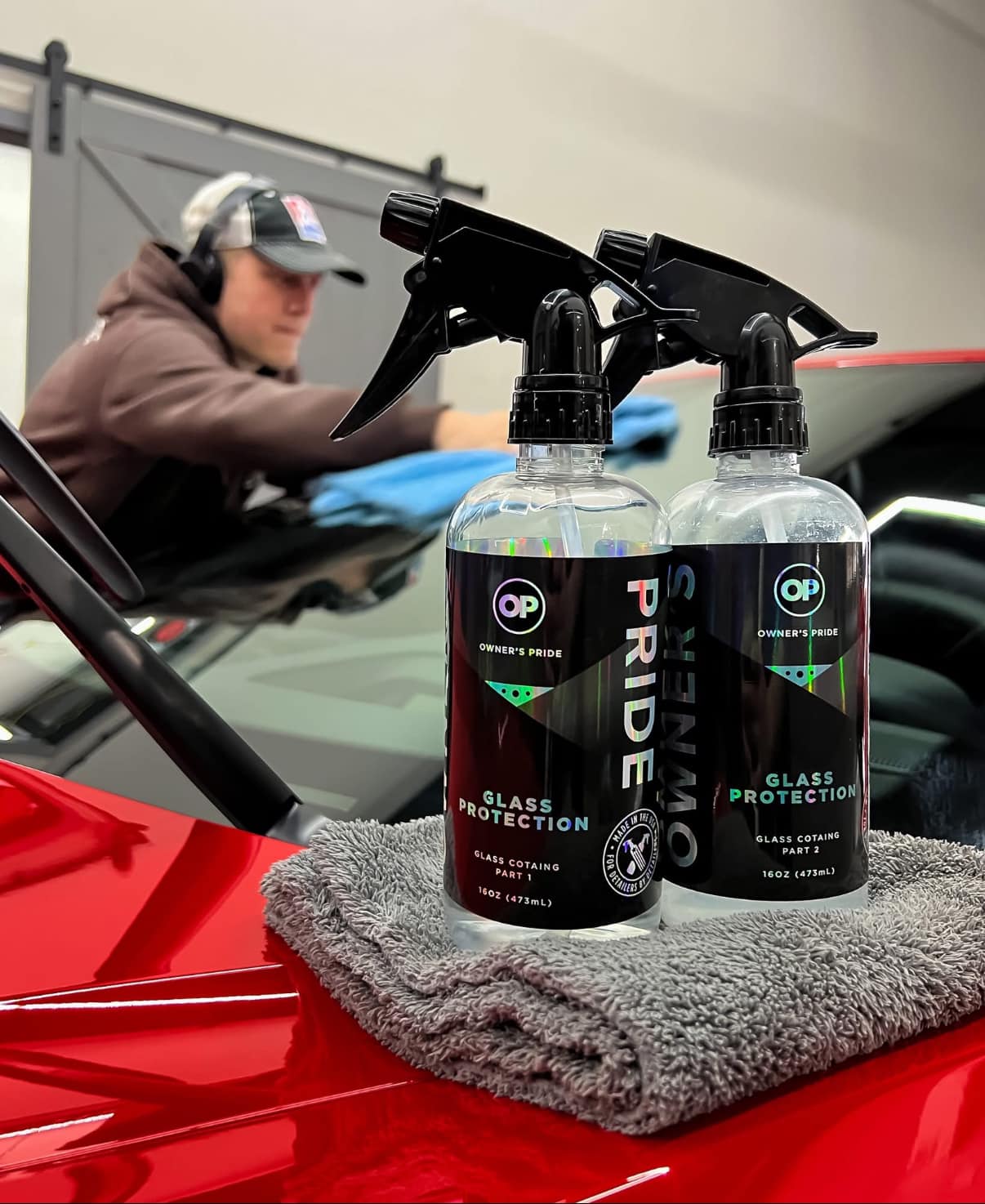 Owner's Pride glass protection products and microfiber towel on a red vehicle, with a technician applying detailing services in the background.
