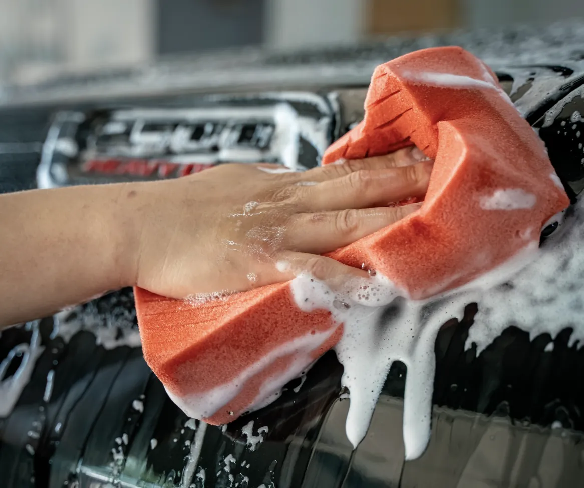 Hand washing a vehicle with an orange sponge, covered in suds, emphasizing the car detailing services offered by Owner's Pride for maintaining ceramic coating and vehicle finish.