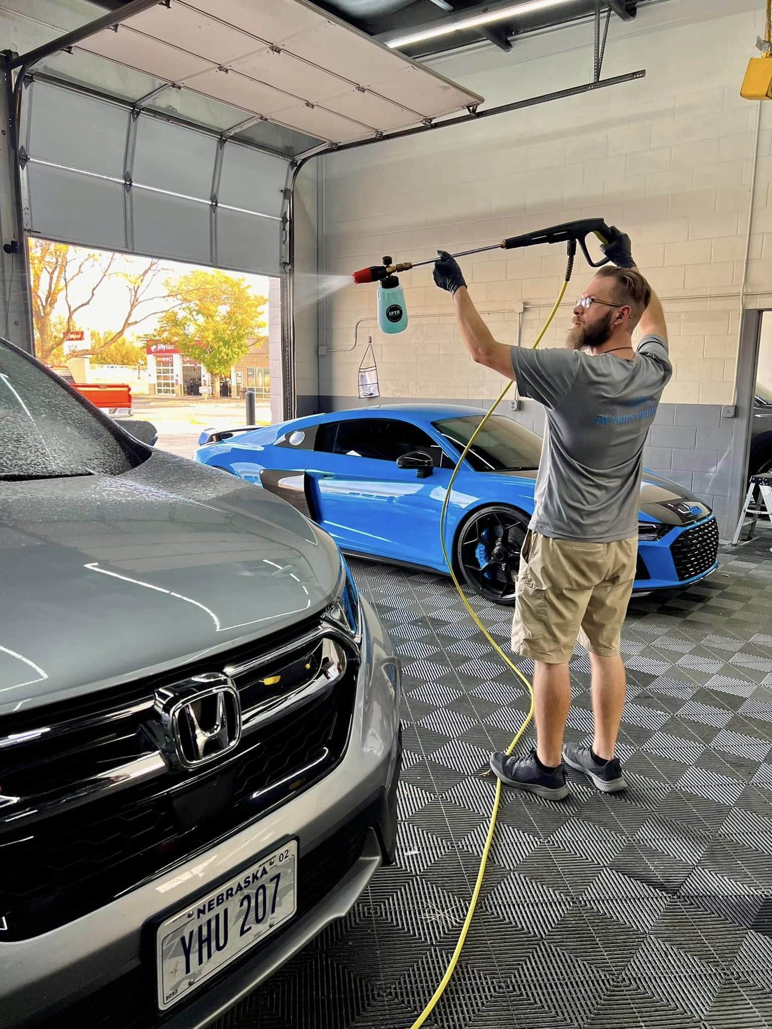 Professional car wash technician spraying vehicle with cleaning solution, showcasing hand wash service in modern car wash facility, with a blue sports car in the background.